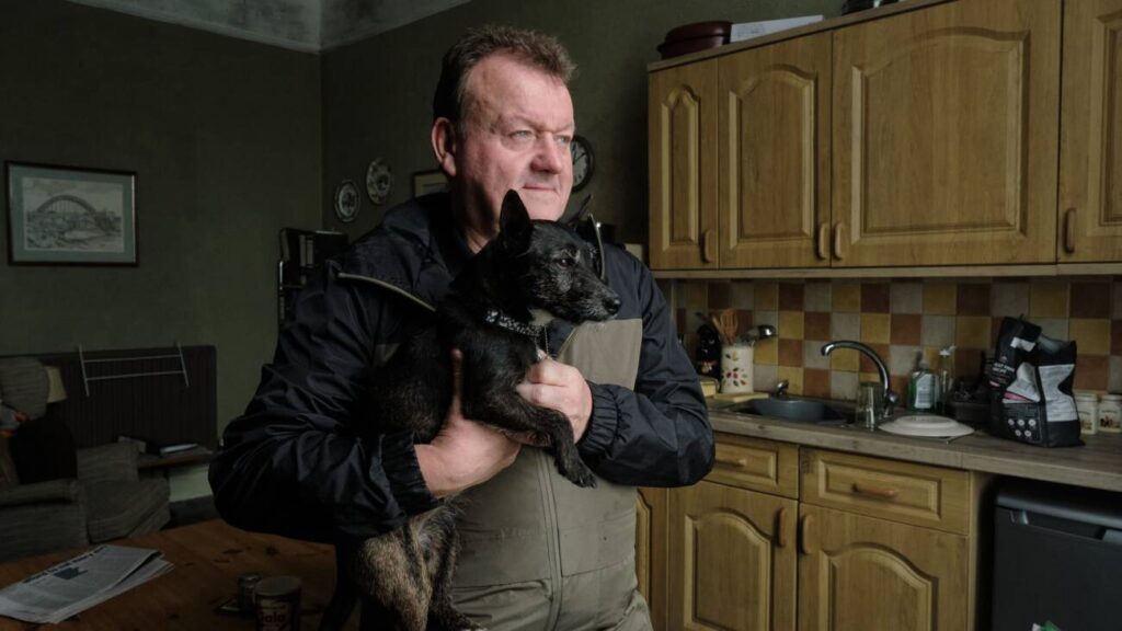 Homem segurando cachorro, em uma cozinha simples.